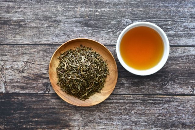 Feuilles de hojicha dans une coupelle en bois à côté d’une tasse de hojicha infusé